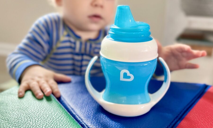 A white baby reaches for a blue Munchkin brand sippy cup on a rainbow foam toy