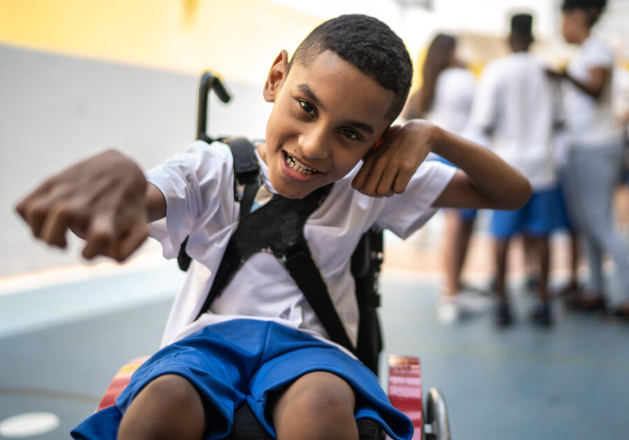 Black young boy with a white t-shirt and blue shorts sits in a wheelchair and smiles