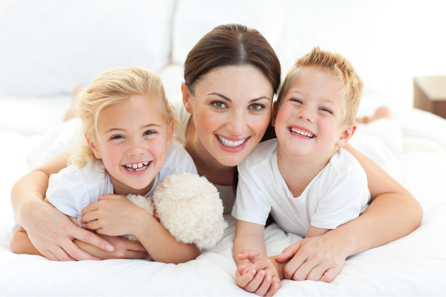 Brunette mom smiles on the bed with her two children, one who has crooked teeth and may need braces