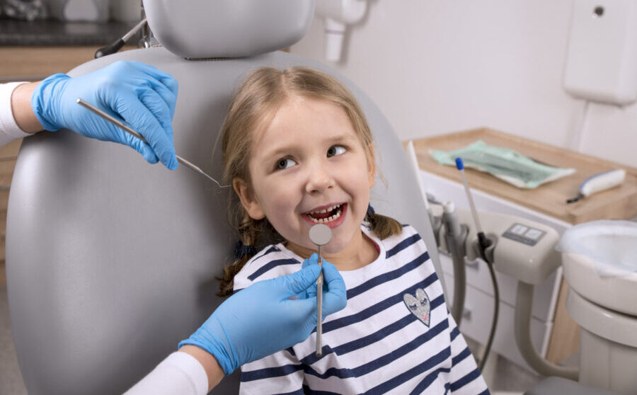 A young girl in a striped shirt sits in a dental chair as her pediatric dentist examines her teeth with special dental tools