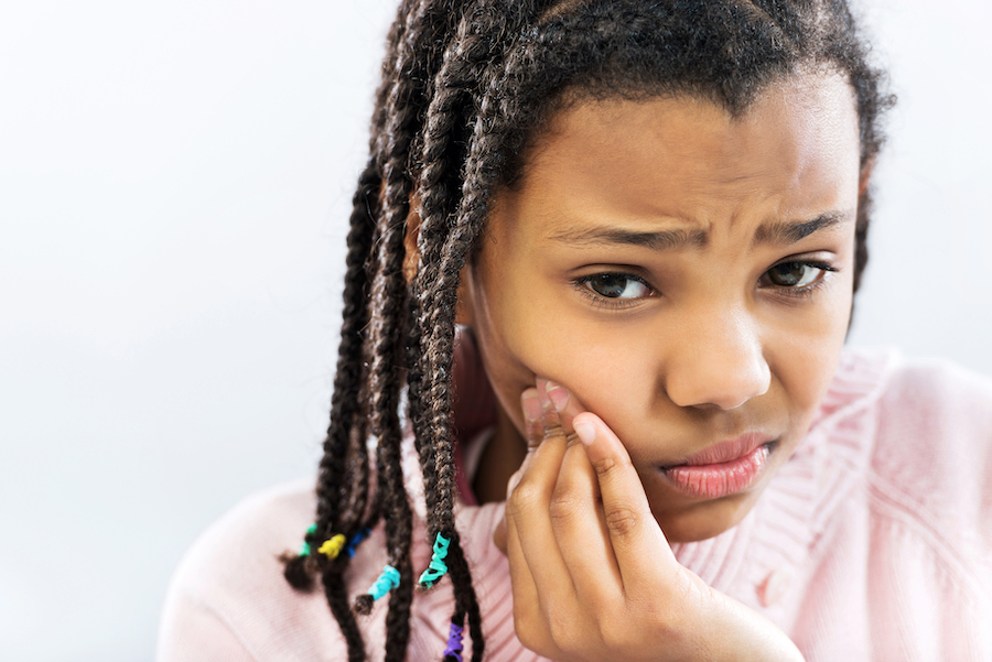 Closeup of a young Black girl cringing in pain and touching her cheek due to tooth pain