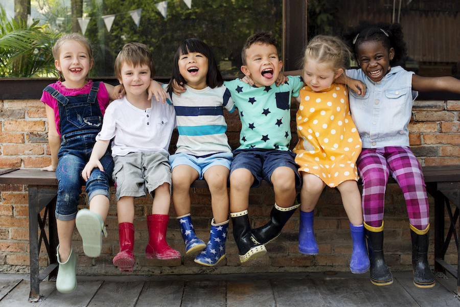 A group of elementary-age children smile while sitting on a bench together wearing rain boots