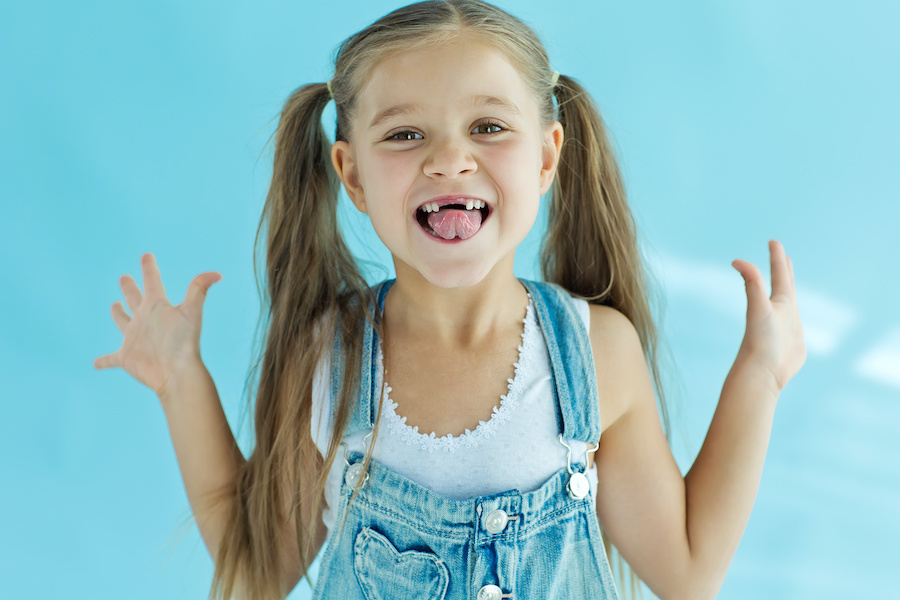 Young girl with pigtails and overalls smiles to show off her missing front teeth after losing her baby teeth
