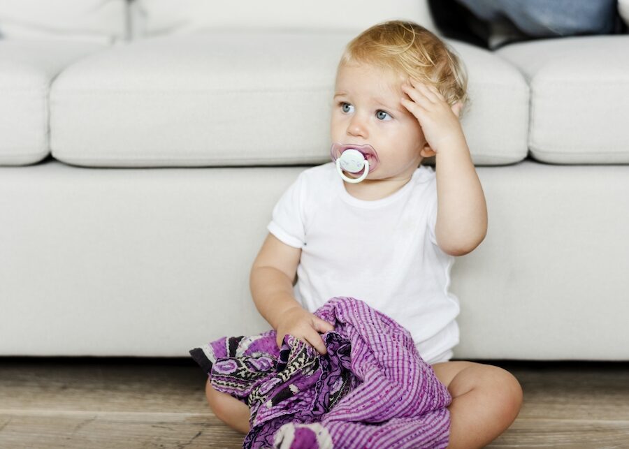 Baby with a pacifier and a  purple blanket sitting on the floor in front of a white couch